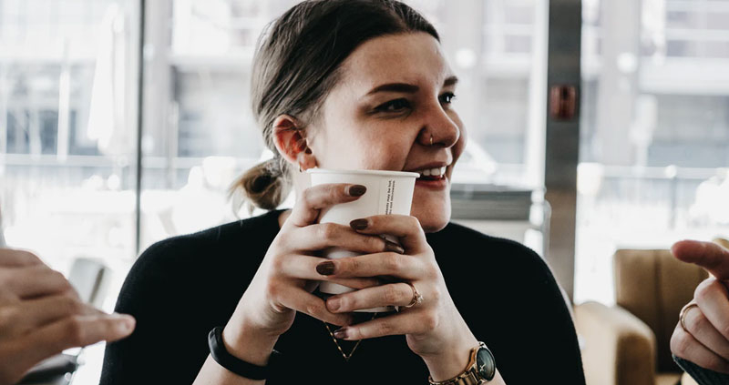 A young woman conversing with two people in a caffe, holding a cup of coffee